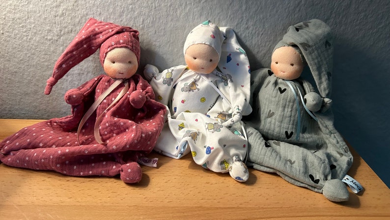 Pu&ograve; includere: Tre bambole di stoffa fatte a mano con volti ricamati e vestiti in tessuto. Le bambole indossano abiti rosa, bianchi e grigi con diversi motivi.