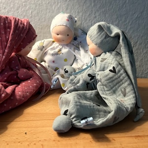 Pu&ograve; includere: Tre bambole di stoffa fatte a mano con visi ricamati e vestiti in tessuto. Le bambole indossano abiti rosa, bianchi e grigi con diversi motivi. Le bambole sono sedute su una superficie di legno.