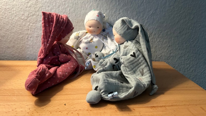 Pu&ograve; includere: Tre bambole di stoffa fatte a mano con visi ricamati e vestiti in tessuto. Le bambole indossano abiti rosa, bianchi e grigi con diversi motivi. Le bambole sono sedute su una superficie di legno.