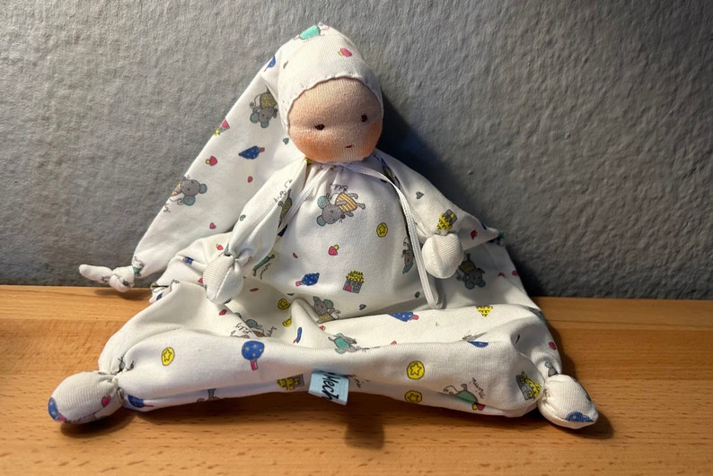 Pu&ograve; includere: Una bambola di stoffa bianca con un cappello di stoffa bianca, che indossa un pigiama bianco e blu a fantasia con un piccolo disegno di topo. La bambola &egrave; seduta su una coperta bianca e blu a fantasia con un piccolo disegno di topo.