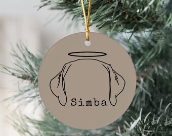 Puede incluir: Adorno navideño redondo de cerámica beige con una cuerda dorada para colgar. Presenta un dibujo lineal negro de la cabeza de un perro con un halo y el nombre "Simba". El adorno cuelga frente a ramas verdes de árbol de Navidad.