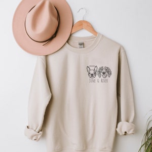 May include: A beige sweatshirt with a crew neck and long sleeves. The sweatshirt features a black and white illustration of two dogs and the text "JUNE & RIVER" below the dog illustrations. A pink hat hangs on a wooden hanger above the sweatshirt.