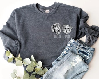 May include: A dark gray sweatshirt featuring two dog portraits with the text "CHARLIE & CAMI" underneath. The sweatshirt is paired with light blue denim jeans and a scrunchie. A sprig of greenery is placed near the clothing.