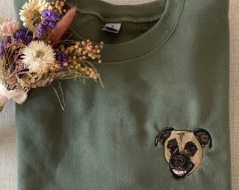 Puede incluir: Una sudadera verde oscuro con una imagen bordada de la cara de un perro. El perro tiene pelo marrón y una mancha blanca en el pecho.