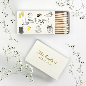 May include: White matchbox with a black and white illustration of a cat, a lemon, a cake, and a dog. The text "Bree & Mike" is written on a black ribbon. The matchbox is closed and the top of the box has a gold inscription that reads "The Kaplans Est. 09.01.25".