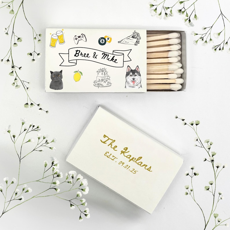 May include: White matchbox with a black and white illustration of a cat, a lemon, a cake, and a dog. The text "Bree & Mike" is written on a black ribbon. The matchbox is closed and the top of the box has a gold inscription that reads "The Kaplans Est. 09.01.25".