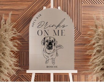 Op de afbeelding: Een bruine en witte hond met de tekst "Drinks on me" en "Open Bar" op een spiegelbord met de naam "Roscoe" onderaan.