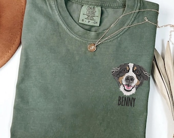 May include: An olive green Comfort Colors t-shirt with a dog face embroidered on the front. The dog has black, brown, and white fur, with the name "Benny" embroidered below. A gold necklace with a pendant rests on the shirt.
