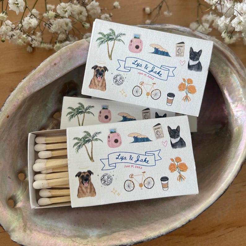 May include: Two white matchboxes with a colorful design featuring a dog, a bicycle, a coffee cup, a palm tree, a wave, and a camera. The matchboxes have a banner that says "Lya & Jake" and "June 11, 2022".