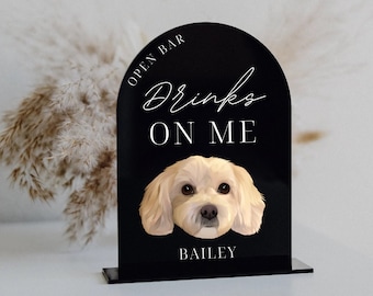 Op de afbeelding: Zwart boogvormig bord met witte tekst die "Open Bar Drinks On Me Bailey" luidt. Een witte hond met bruine ogen is afgebeeld op het bord.