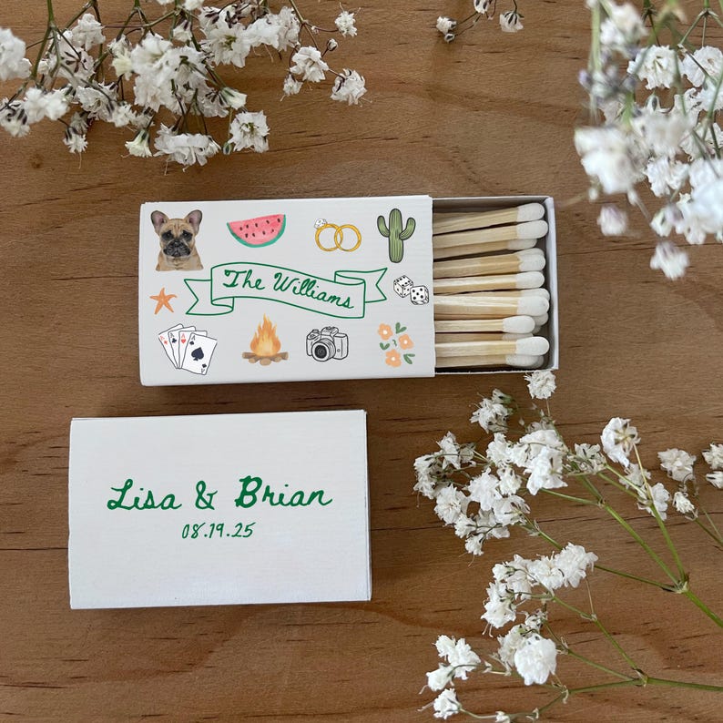 May include: Two white matchboxes with green text. The top box has a colorful illustration of a dog, watermelon, rings, cactus, dice, playing cards, a campfire, and a camera. The text reads "The Williams". The bottom box has the text "Lisa & Brian 08.19.25".