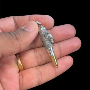 May include: Silver-toned bullet pendant with a gold-colored tip. The pendant features a detailed, textured design around the bullet's body and a small loop for a chain. The background is solid black.