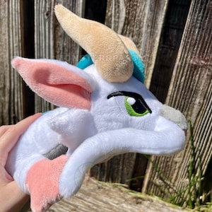 May include: A plush white creature with pink and blue accents, featuring large, expressive eyes with green irises and black outlines. The plush has tan horns and pink inner ears, creating a whimsical design.