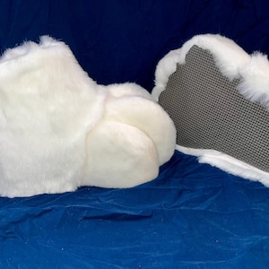 May include: Pair of white furry costume feet, likely for a mascot or character. The feet are plush and fluffy, with a textured gray sole. The fur is a bright white, and the overall shape suggests a large animal foot.