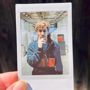 Può includere: Una fotografia istantanea quadrata che ritrae una persona con capelli castano chiaro che indossa un maglione blu scuro con mezza zip. La persona ha una mano appoggiata al mento. Lo sfondo mostra un lungo corridoio industriale con strutture bianche.