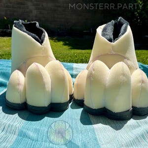 May include: A pair of cream-colored, foam-based monster feet with black soles and dark gray interior lining. The feet have large, rounded toes and are designed to be worn. The text "MONSTERR.PARTY" is visible in the upper right corner.