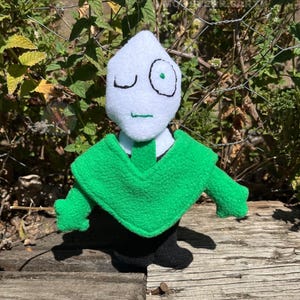 May include: A handmade plush doll with a white head, winking eye, and green eye. The doll wears a green cape and tie over a white shirt, with black pants. The doll is posed on a wooden surface with a natural background.
