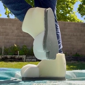 May include: A pair of oversized, off-white foam shoes with a textured gray sole. The shoes are worn by a person wearing blue jeans. The shoes are designed to look like cartoon feet.