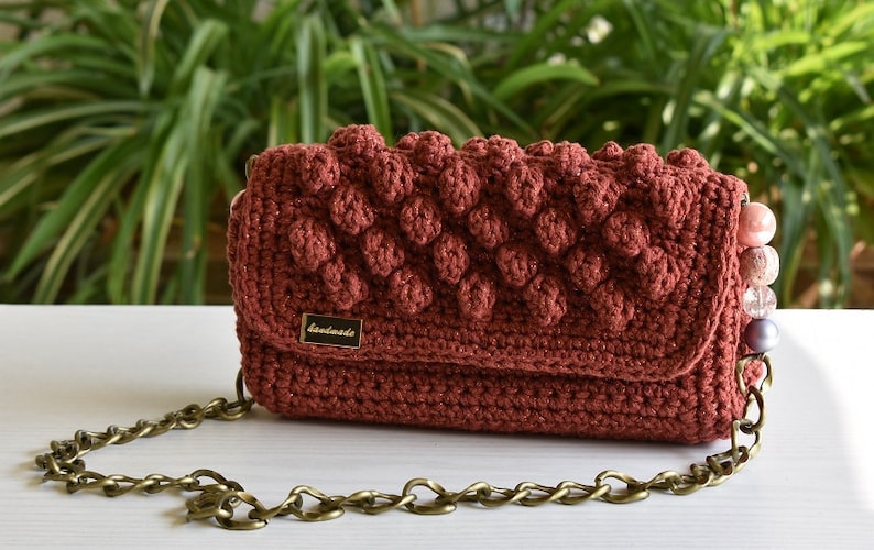 Op de afbeelding: Een bordeauxrode gebreide handtas met een kettingband en een gouden label met de tekst "handgemaakt".