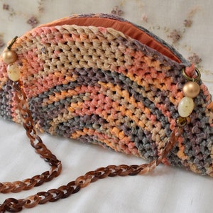 Puede incluir: Un bolso de mano de crochet en forma de semic&iacute;rculo con una correa de cadena marr&oacute;n y cuentas decorativas. El bolso est&aacute; hecho con hilo multicolor en tonos naranja, rosa y gris.