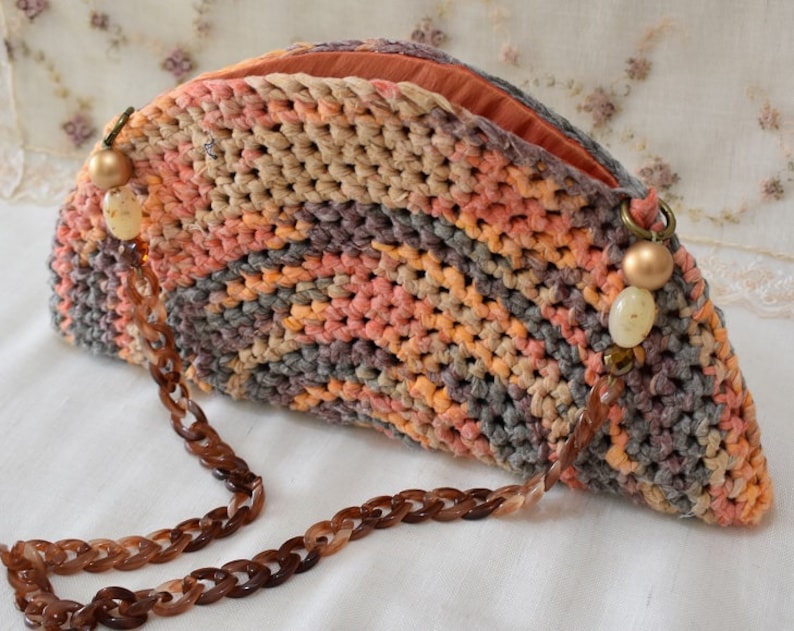Puede incluir: Un bolso de mano de crochet en forma de semic&iacute;rculo con una correa de cadena marr&oacute;n y cuentas decorativas. El bolso est&aacute; hecho con hilo multicolor en tonos naranja, rosa y gris.