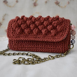Op de afbeelding: Een klein, rood gehaakt handtasje met een kettingband en parel accenten. De tas heeft een textuur, een bol design.