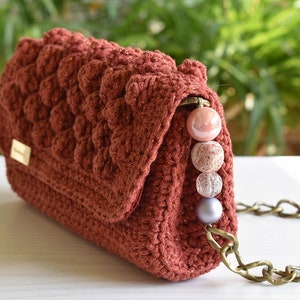 Op de afbeelding: Een roestkleurige gehaakte handtas met een kettingband en een rij roze en transparante kralen.