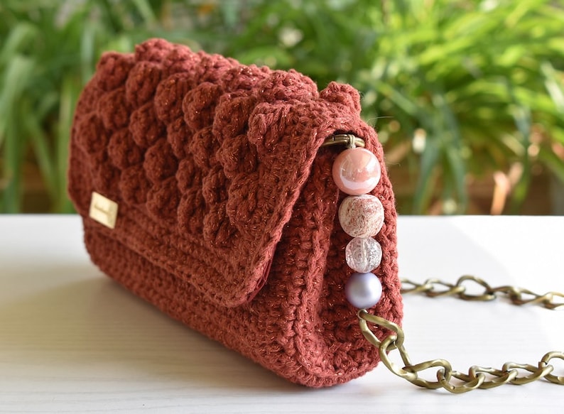 Op de afbeelding: Een roestkleurige gehaakte handtas met een kettingband en een rij roze en transparante kralen.