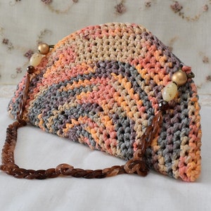 Puede incluir: Un bolso de mano de crochet con forma de semic&iacute;rculo, con un patr&oacute;n multicolor de marr&oacute;n, naranja y gris. El bolso tiene una correa de cadena marr&oacute;n con cuentas decorativas.