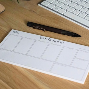 Könnte beinhalten: Ein weißer Wochenplaner mit dem deutschen Wort "Wochenplan" aufgedruckt, zusammen mit dem Text "Nice & Noted". Ein dunkler Stift liegt auf dem Planer, in der Nähe einer Computertastatur auf einer Holzoberfläche.
