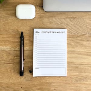 Op de afbeelding: Een witte notitieblok met de tekst "Eins nach dem Anderen" en een pen op een houten bureau. Het notitieblok heeft een lijstformaat met selectievakjes. Een witte draadloze oortjes case ligt ook op het bureau.