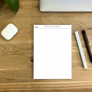 Puede incluir: Un bloc de notas blanco con cuadrícula de puntos con el texto "Platz für Geistesblitze" está sobre un escritorio de madera. Un bolígrafo blanco, un bolígrafo negro y una funda blanca para auriculares inalámbricos también están sobre el escritorio. El bloc de notas es de la marca "Nice & Noted".