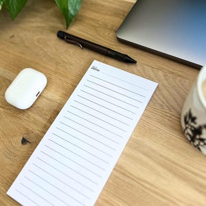 Könnte beinhalten: Ein weißer, liniierter Notizblock mit dem Text "Yet Noted" liegt auf einem Holzschreibtisch, neben einem Stift, einem weißen kabellosen Ohrhörer-Etui, einem Laptop und einer Kaffeetasse. Der Notizblock ist bereit für Notizen.