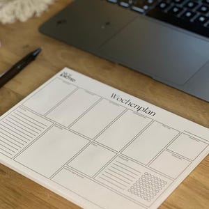 Könnte beinhalten: Ein weißer Wochenplaner-Block mit minimalistischem Design. Der Block hat Abschnitte für jeden Wochentag, Notizen und Tracking. Der Text "NOTED" und "Wochenplan" sind auf dem Block aufgedruckt. Ein Laptop und ein Stift befinden sich im Hintergrund.