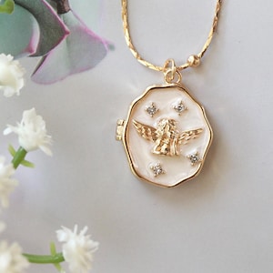 May include: A gold-colored necklace with a pendant featuring an angel design. The pendant is round with a white enamel background and gold trim. The angel is gold-colored with outstretched wings. The necklace chain has small gold beads and a delicate design. The background includes white flowers and a soft, neutral color palette.