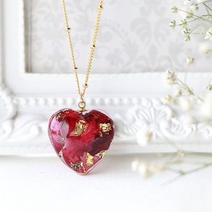 May include: A gold chain necklace with a heart-shaped pendant filled with red rose petals and gold flakes.