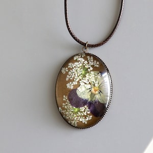 May include: A brown cord necklace with an oval pendant featuring a pressed flower design. The pendant has a clear resin background and a bronze-colored metal frame. The flower design includes a white flower with a purple center and small white flowers.