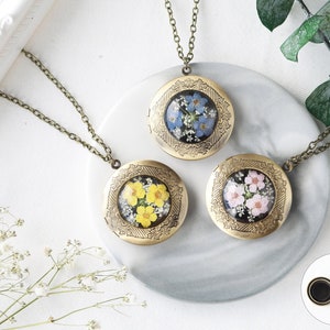 May include: Three gold locket necklaces with different pressed flower designs. The lockets are round and have a detailed floral border. The flowers are blue, yellow, and pink.