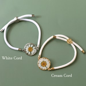 May include: Two daisy flower bracelets with adjustable white and cream cords. One bracelet has a silver-tone setting, and the other has a gold-tone setting. The text "White Cord" and "Cream Cord" are visible.
