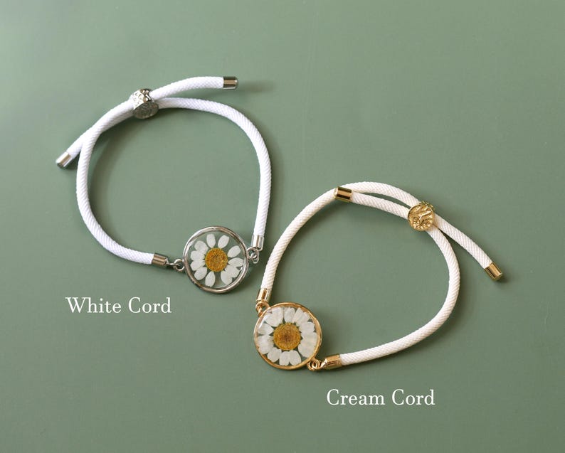 May include: Two daisy flower bracelets with adjustable white and cream cords. One bracelet has a silver-tone setting, and the other has a gold-tone setting. The text "White Cord" and "Cream Cord" are visible.