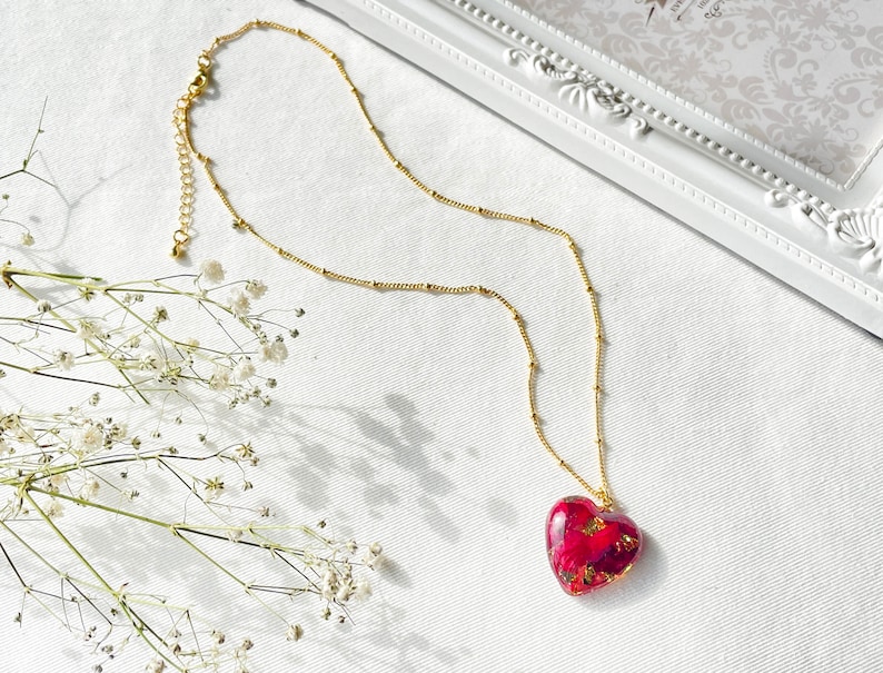K&ouml;nnte beinhalten: Eine goldene Kettenkette mit einem herzf&ouml;rmigen Anh&auml;nger, der mit roten und goldenen gepressten Blumen gef&uuml;llt ist.