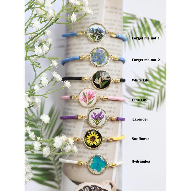 May include: A collection of seven bracelets with gold-colored metal settings and colorful cords. Each bracelet features a different pressed flower, including forget-me-nots, lilies, lavender, a sunflower, and a hydrangea. The text on the right reads: Forget me not 1, Forget me not 2, White Lily, Pink Lily, Lavender, Sunflower, Hydrangea.
