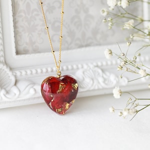 K&ouml;nnte beinhalten: Eine goldene Kettenkette mit einem herzf&ouml;rmigen Anh&auml;nger, gef&uuml;llt mit roten Rosenbl&auml;ttern und Blattgold.