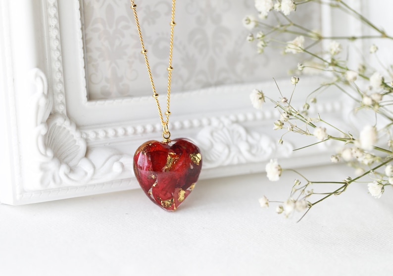 K&ouml;nnte beinhalten: Eine goldene Kettenkette mit einem herzf&ouml;rmigen Anh&auml;nger, gef&uuml;llt mit roten Rosenbl&auml;ttern und Blattgold.