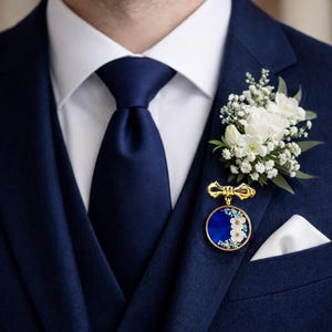 May include: A navy blue suit with a white dress shirt and a navy blue tie. A gold-toned brooch with a blue and white floral design is pinned to the lapel, complemented by a white floral boutonniere and a white pocket square.