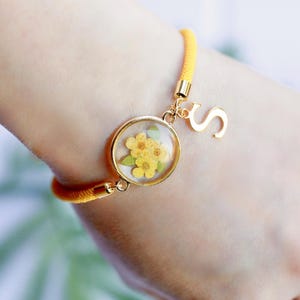 May include: Yellow cord bracelet with a gold-colored charm featuring dried yellow flowers encased in a clear resin. A gold letter "S" charm hangs next to the floral charm. The bracelet is on a wrist.