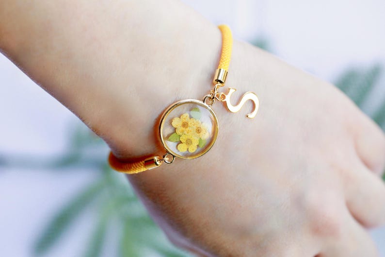 May include: Yellow cord bracelet with a gold-colored charm featuring dried yellow flowers encased in a clear resin. A gold letter "S" charm hangs next to the floral charm. The bracelet is on a wrist.