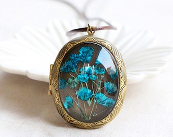 May include: An oval antique bronze locket pendant with a clear glass front, displaying dried blue flowers. The pendant hangs from a brown cord. The locket has a decorative, ornate border.