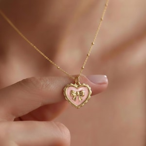 May include: A gold necklace with a heart-shaped pendant. The pendant has a pink enamel fill and a bow design, with a scalloped edge. The chain is delicate with small, evenly spaced beads.