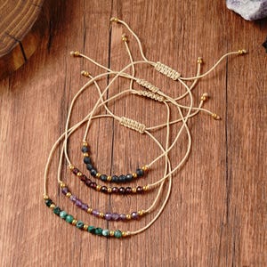 May include: Four beaded bracelets with adjustable beige cords. Each bracelet features a different color of small beads, including black, red, purple, and green, accented with gold-colored beads. The bracelets are displayed on a wooden surface.